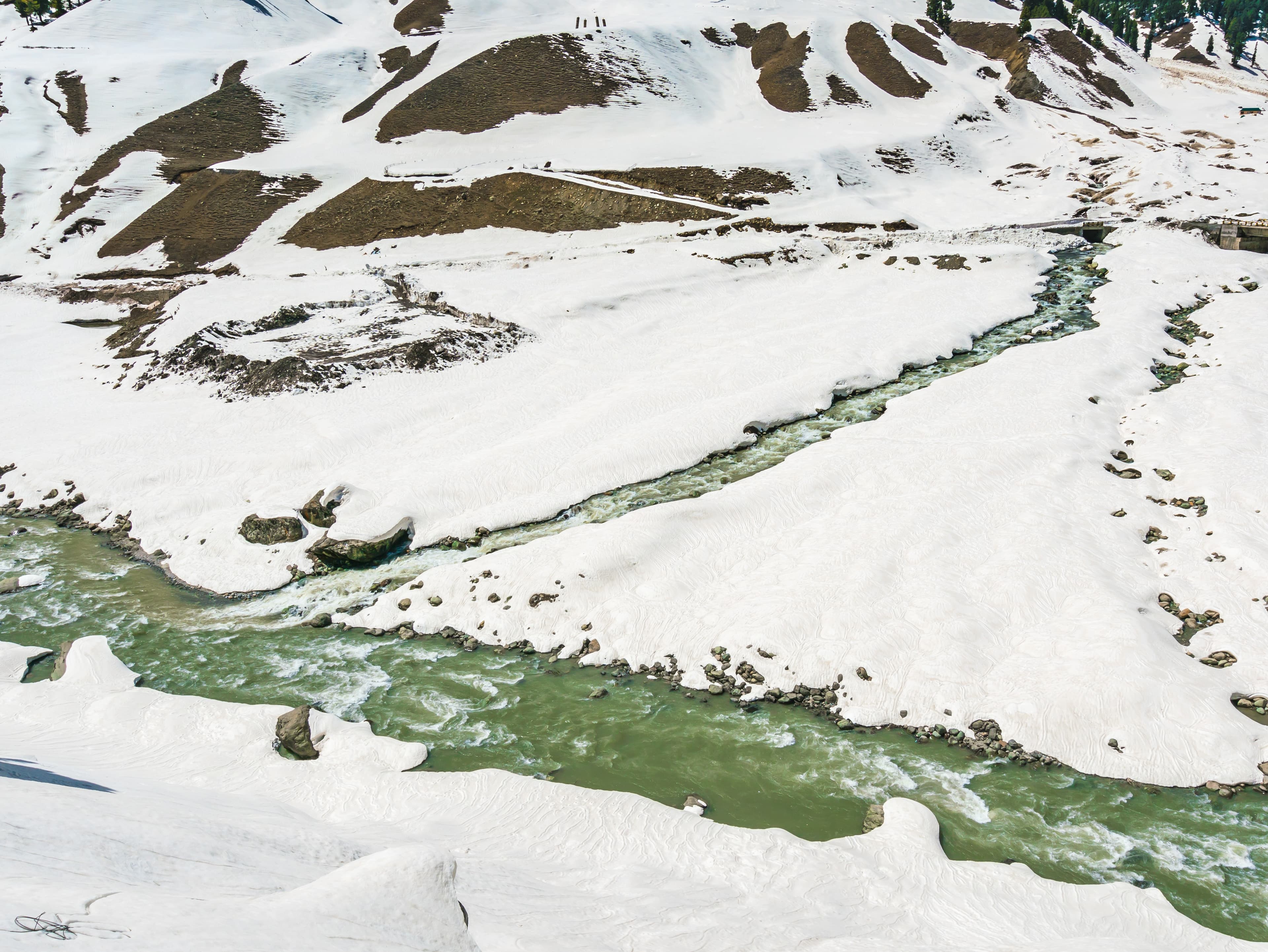 An elevated view of a glacier river flowing elegantly across a broad valley, flanked by snow-covered mountains, demonstrating the stunning beauty and geological elements of a high-altitude Himalayan environment.