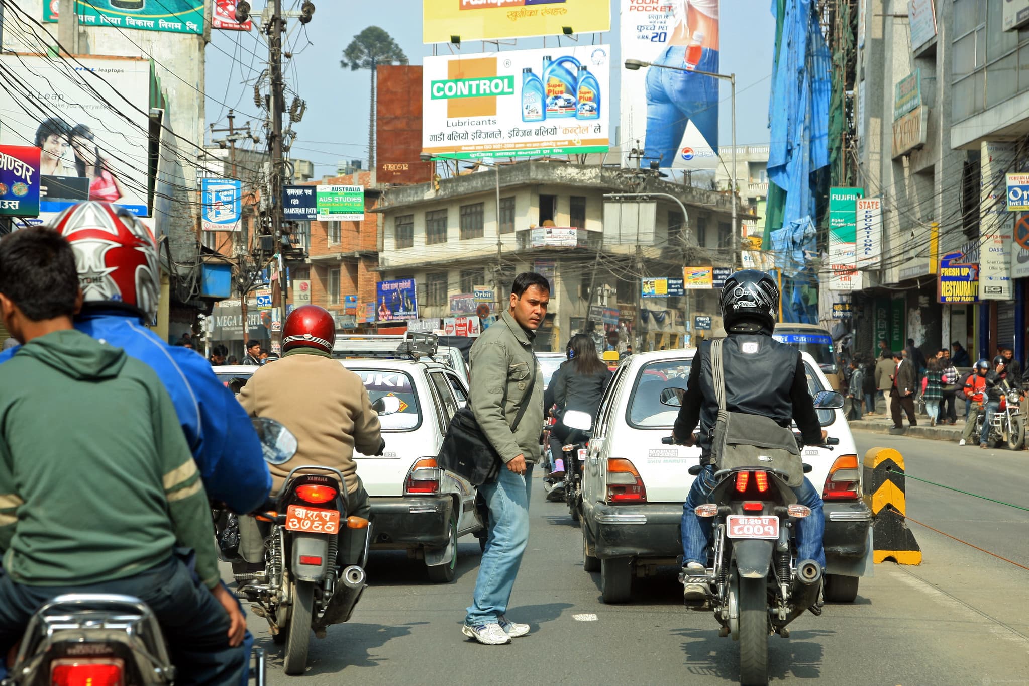 Image of a man in a light jacket and jeans walks across a busy city street, surrounded by motorcycles, cars, and buildings with signs, under a crisscross of overhead wires.